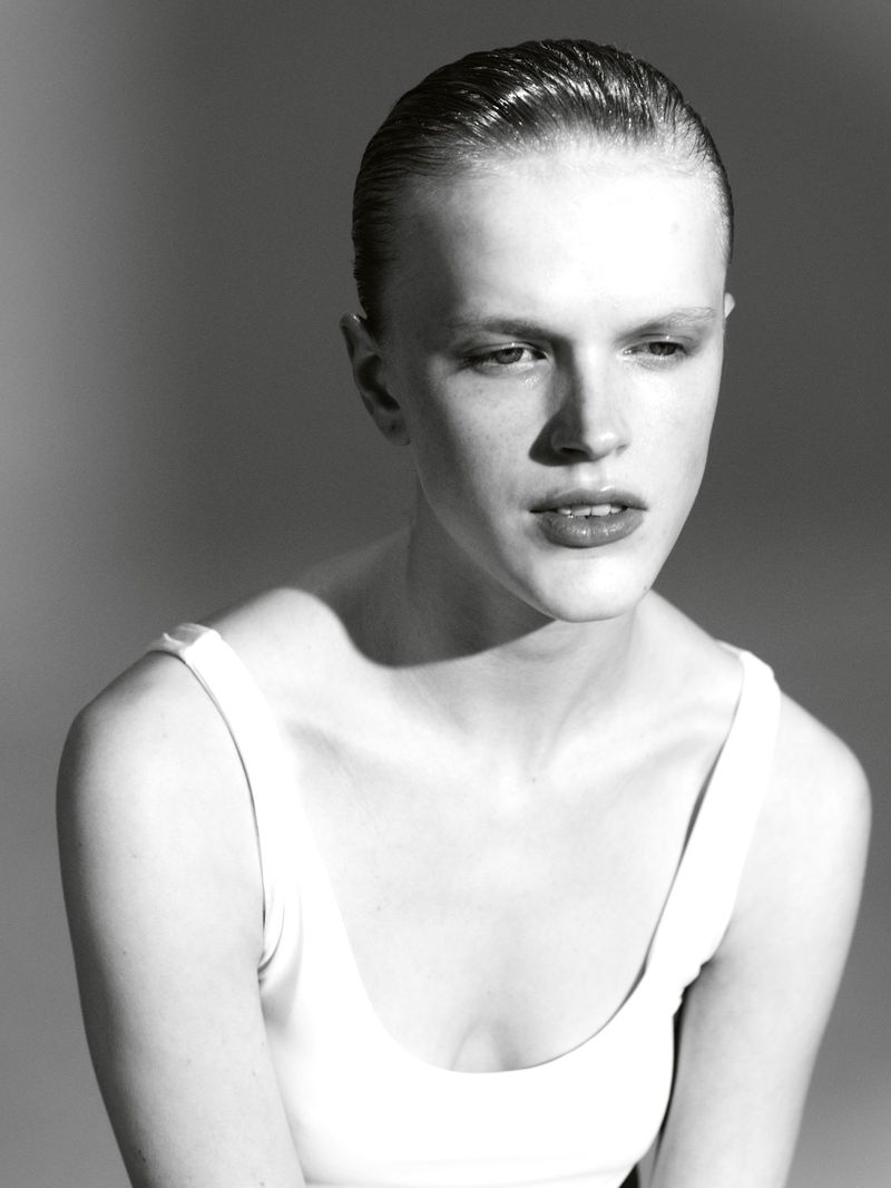 Striking black and white studio portrait of a young adult female with slicked-back hair, wearing a white tank top against a grey background.