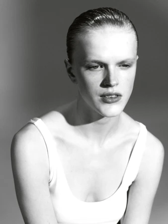 Striking black and white studio portrait of a young adult female with slicked-back hair, wearing a white tank top against a grey background.