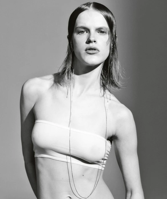 Portrait of Hannah Motler wearing a white strapless top and long delicate chain necklaces against a grey studio backdrop.