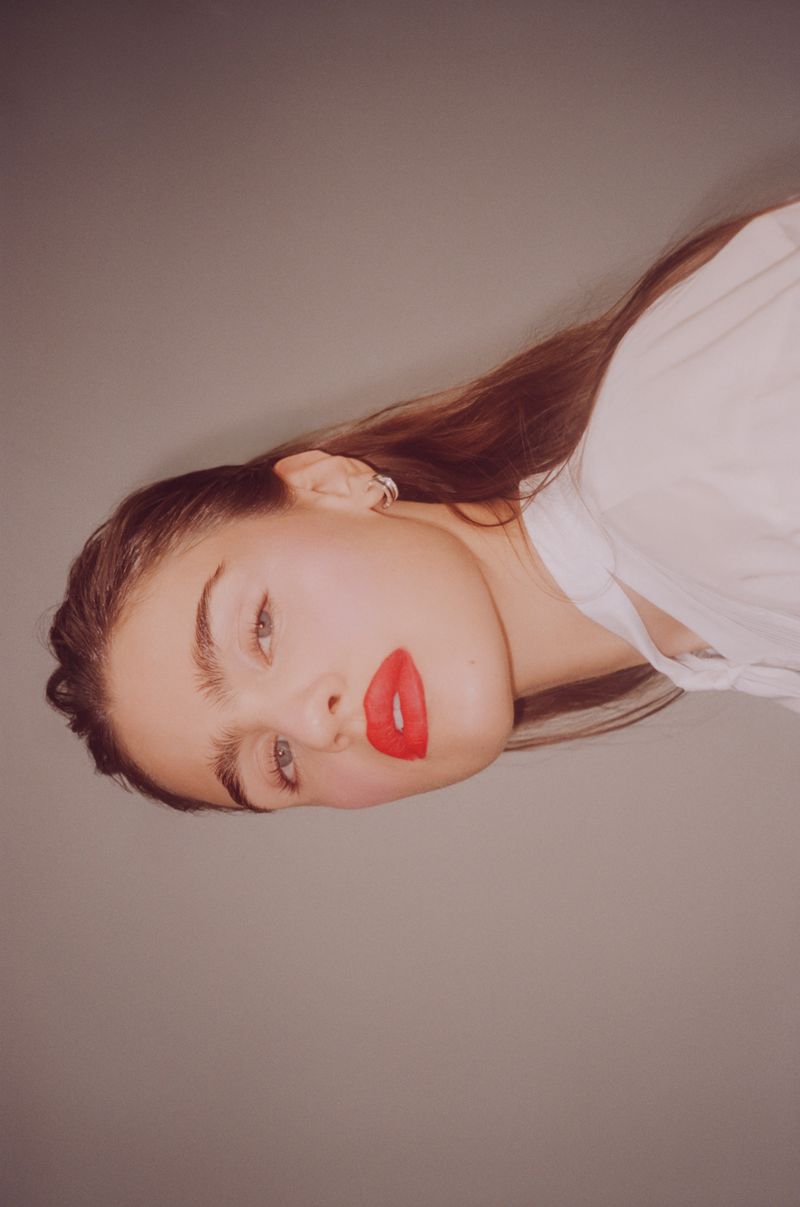 Close-up portrait of a young adult woman with long brown hair, red lipstick, and a white shirt, set against a neutral gray background.