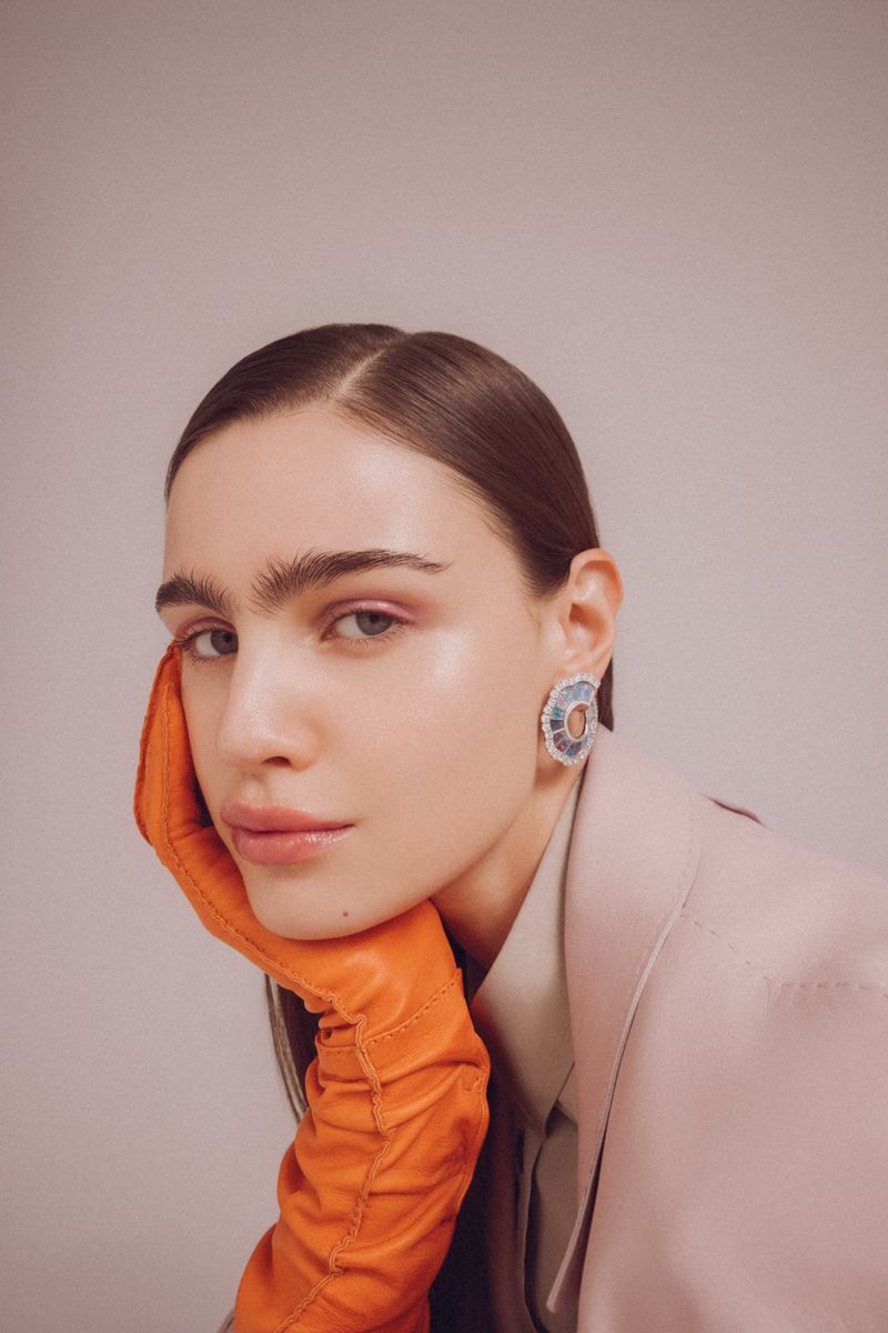 Close-up of a woman with dark hair posing with her face resting on her hand, wearing large earrings, a light jacket, and orange gloves.