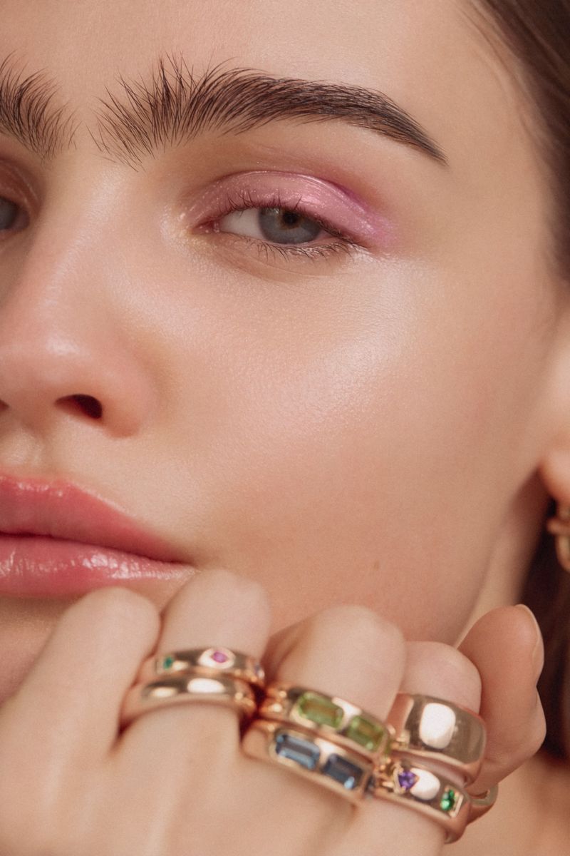 Close-up of a young woman's face with pastel pink eyeshadow and shiny pink lips, wearing a collection of gem-studded gold rings.