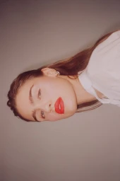 Close-up portrait of a woman with red lipstick and long brown hair wearing a white top against a grey seamless background