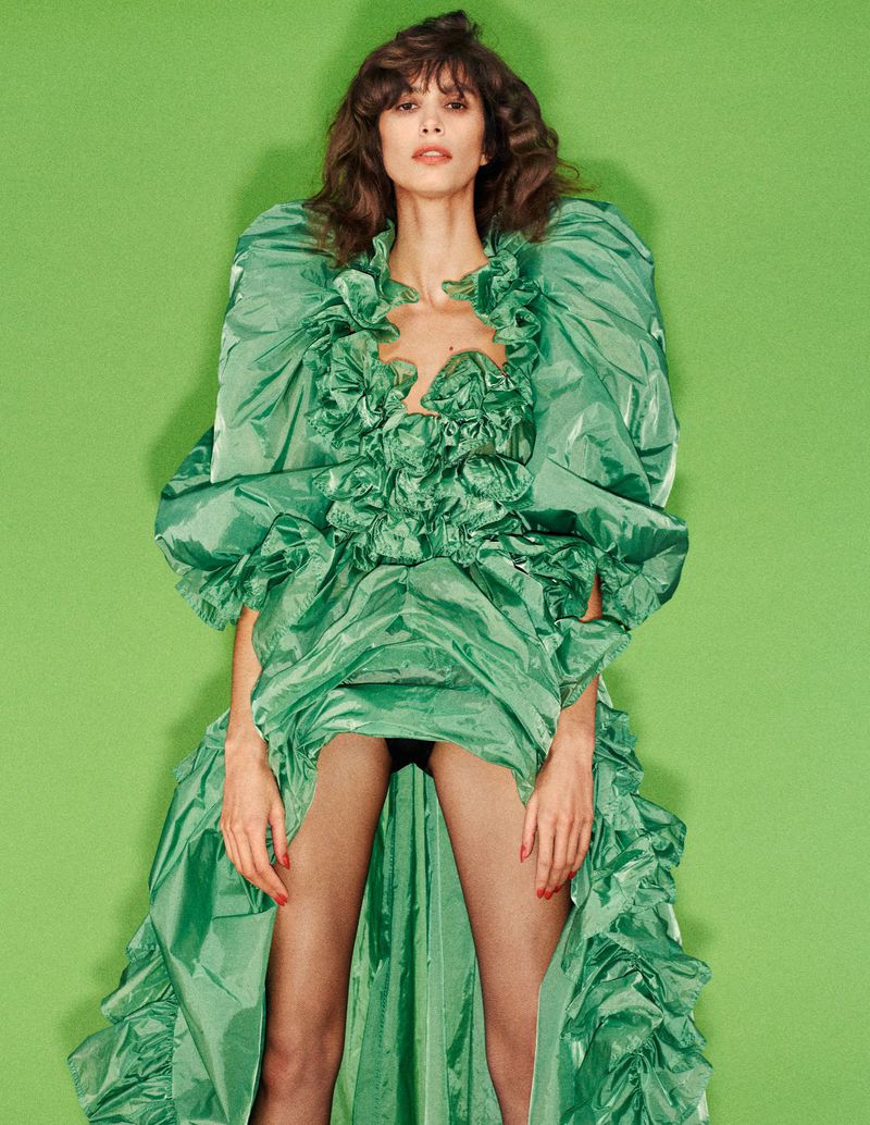 A brunette woman poses in a ruffled green gown against a bright green backdrop, creating a vibrant and eye-catching fashion portrait.