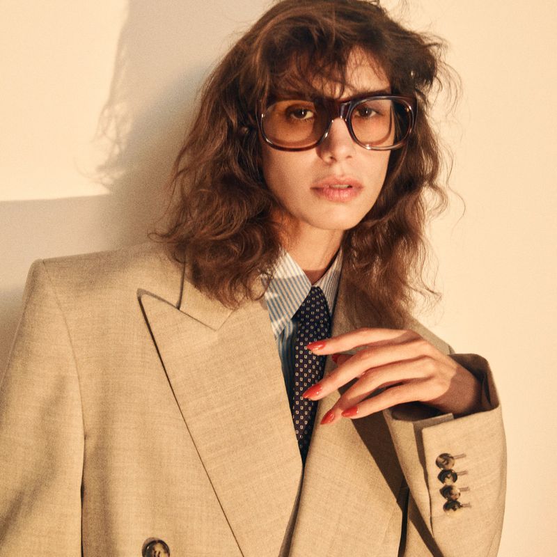 Portrait of a woman with wavy brown hair wearing large, square sunglasses, a beige blazer, striped shirt, and a navy blue tie.