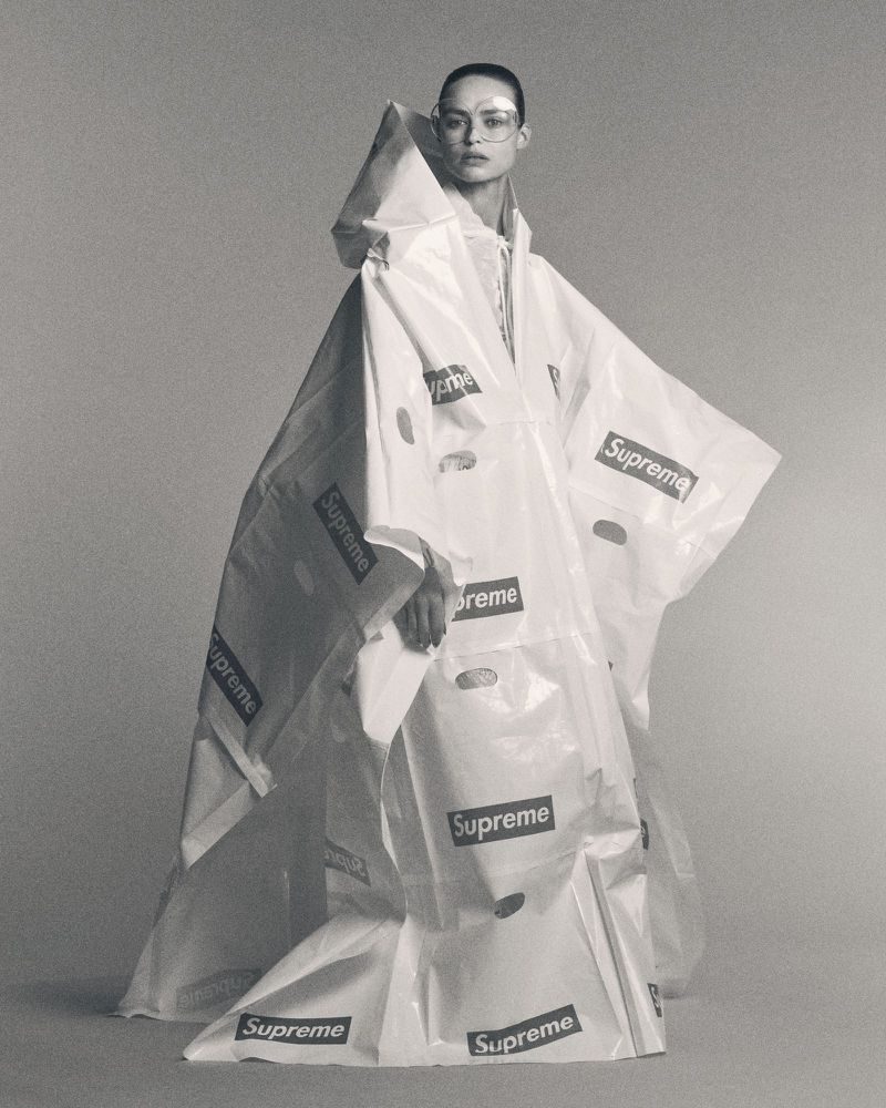 Fashion model poses wearing a hooded cape made from white plastic shopping bags with the brand name Supreme repeated on the bags.