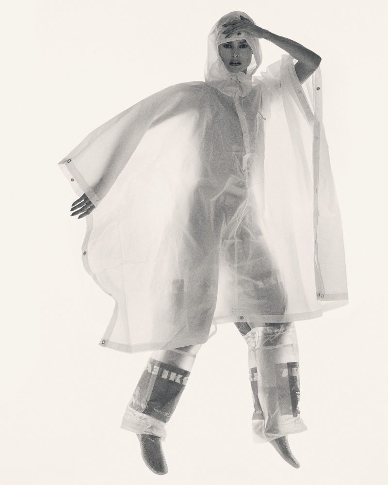 Monochrome fashion shot of an adult woman in translucent raincoat and pants, posing with hand raised in a bright indoor studio.