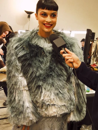 A woman with dark hair and red lipstick being interviewed, wearing a shaggy silver top, indoors.