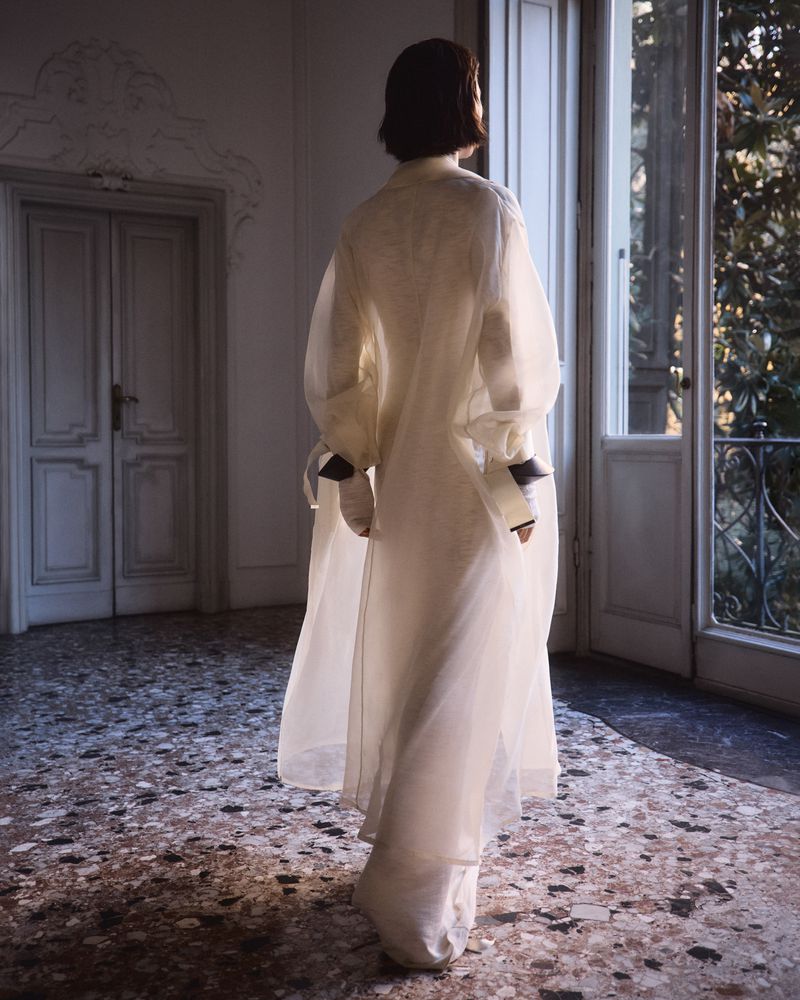 Woman in sheer white dress stands in ornate room with sunlight streaming through windows, showcasing fashion and interior design elements.