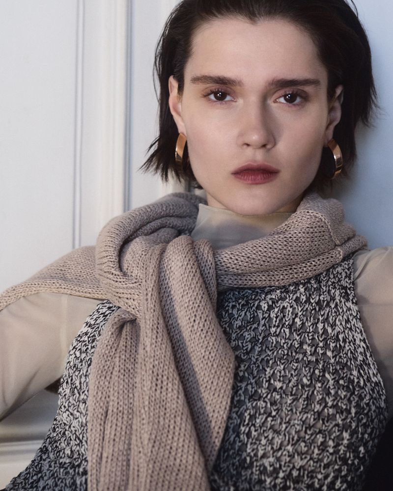 Portrait of a woman wearing a beige scarf, a patterned vest, and gold hoop earrings, posing indoors against a white wall.
