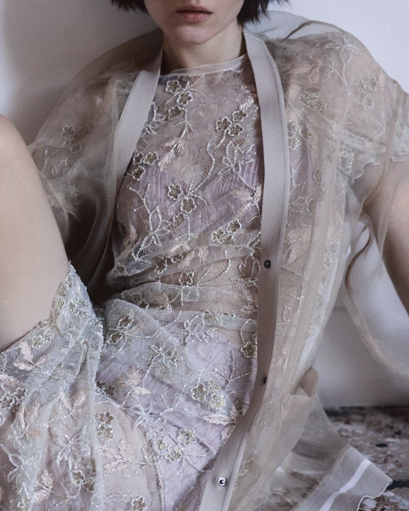 Woman wearing a sheer, embroidered shirt and jacket, sitting indoors against a white wall. Light, airy, elegant fashion image.