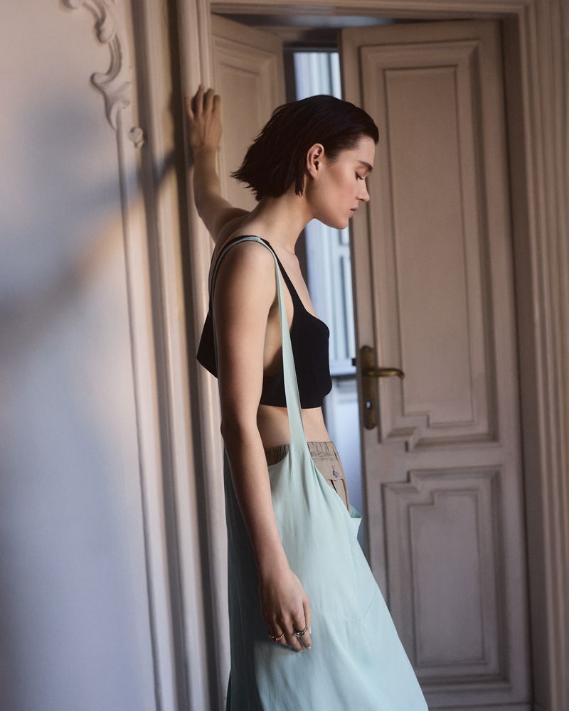 Indoor portrait of a young woman in a black top and skirt, leaning against a wall near an open doorway. Soft, diffused natural light.