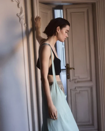 Indoor portrait of a young woman in a black top and skirt, leaning against a wall near an open doorway. Soft, diffused natural light.