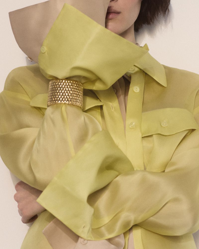 Close-up of a person in a chartreuse collared shirt with a large gold bracelet covering the lower face with a beige garment.