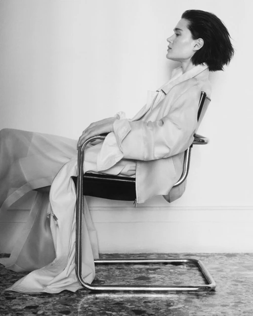 Monochrome fashion portrait of an adult woman in a light coat, seated in a modern chair against a white wall, in a photo studio.