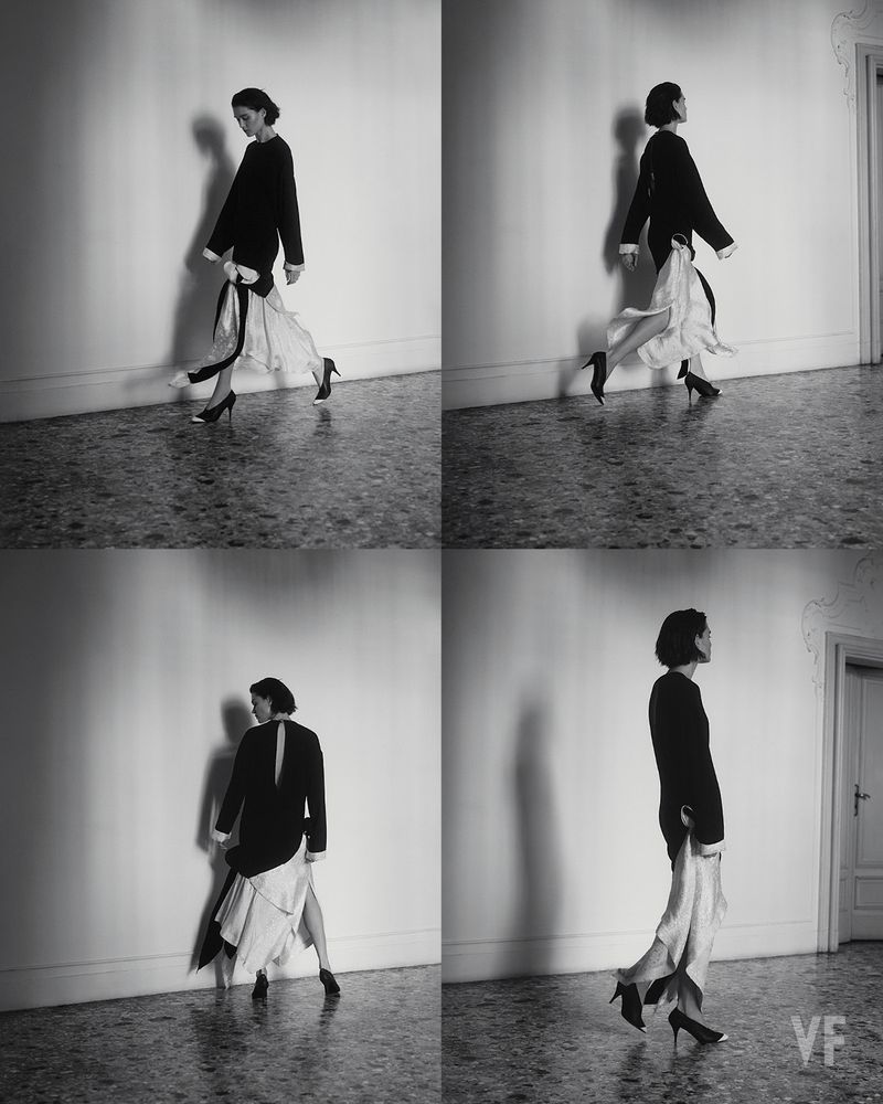 Woman in black top and flowing white skirt walks in a photo collage, with a light wall background, black heels, and indoor setting.