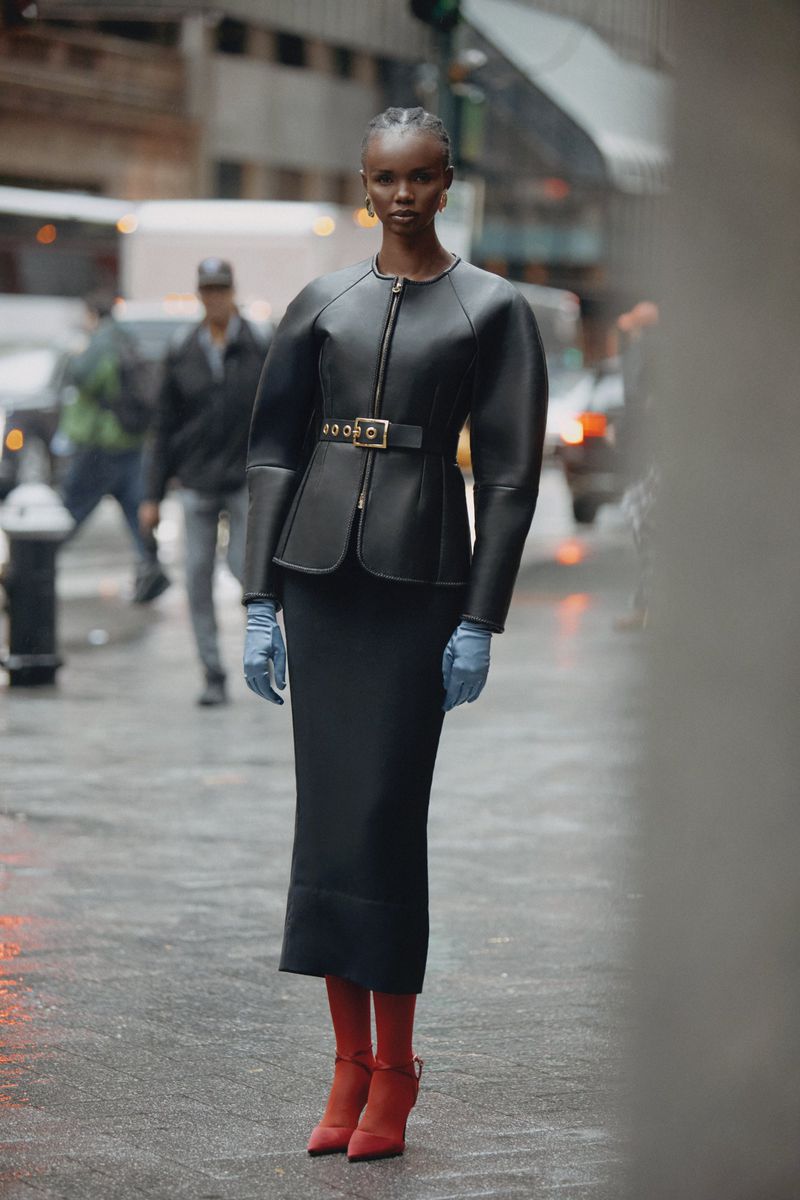 Woman in black jacket and skirt stands on a city street with pedestrians and vehicles in the background; she wears red boots and blue gloves.