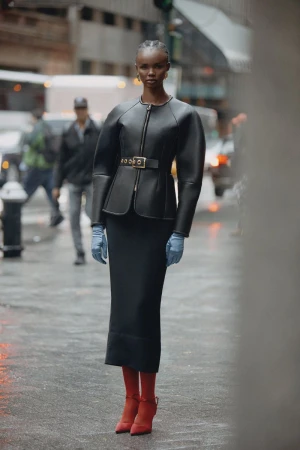 Woman in black jacket and skirt stands on a city street with pedestrians and vehicles in the background; she wears red boots and blue gloves.