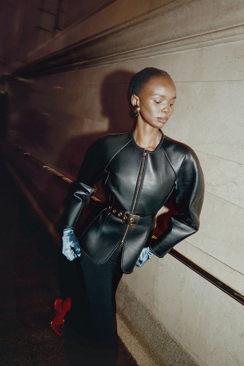 Elegant Black woman in runway fashion poses in an underground tunnel wearing a black leather jacket with blue gloves and red heels.