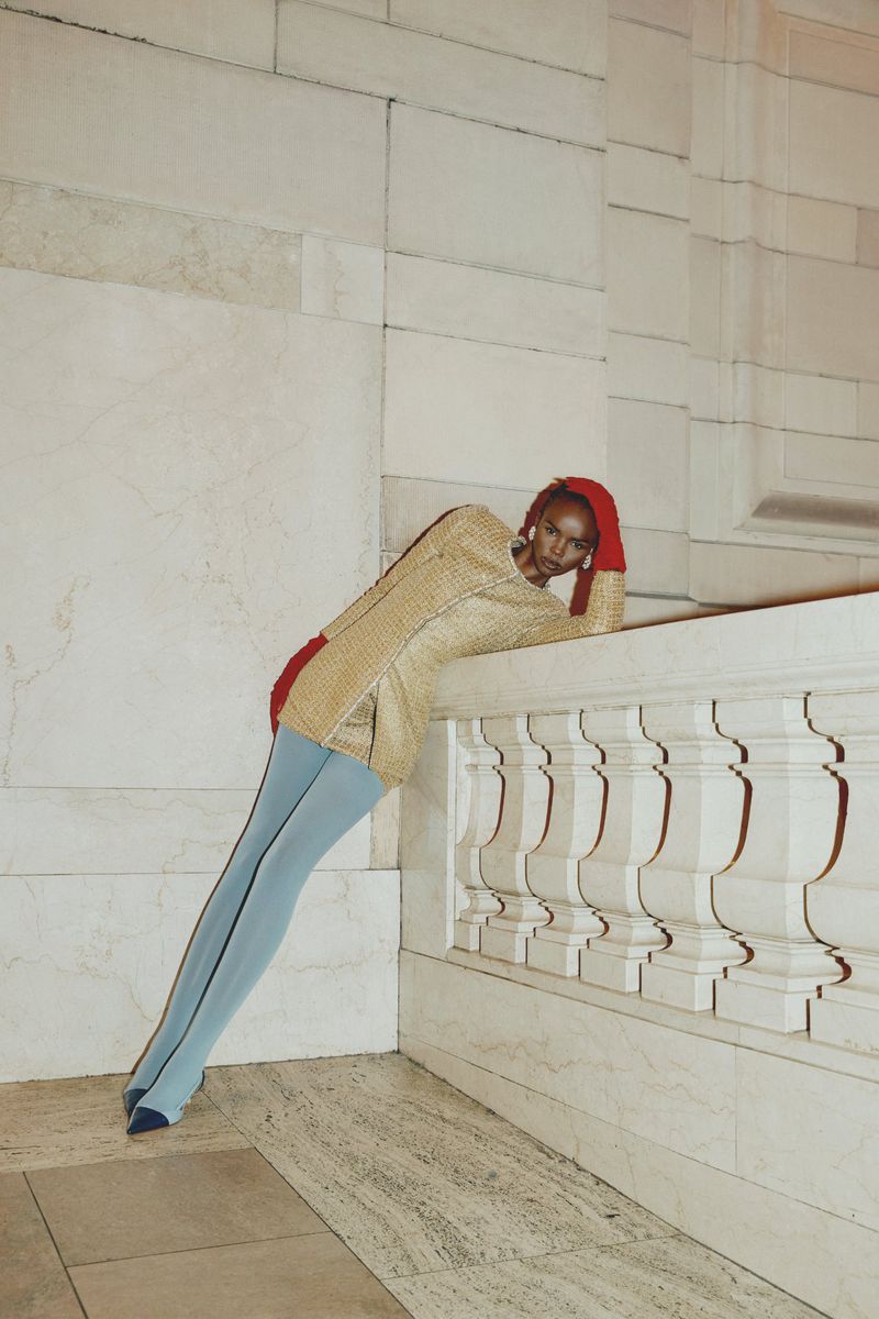 Elegant woman in a gold jacket and light blue tights poses against a marble wall, creating a striking fashion statement with red gloves.