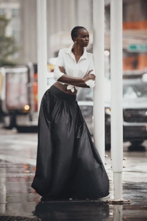 Woman in white shirt and long black skirt leaning against white poles on a wet city street with blurry cars and buildings.