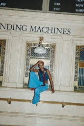 Model wearing blue trousers and a dark top sitting against an ornate stone wall with vending machines sign