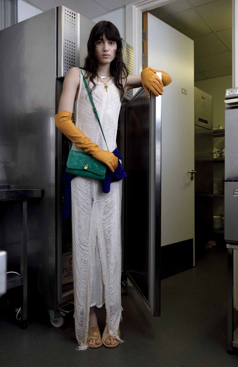 A slender woman in a flowing white dress, accessorized with orange gloves and a green purse, leans against a stainless steel refrigerator.