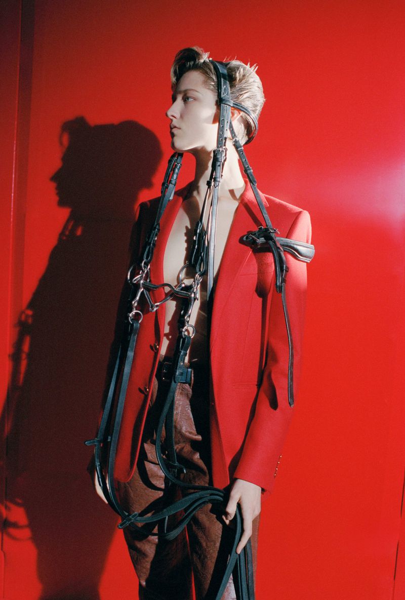 Adult wearing a red blazer and horse bridle stands indoors against a matching red backdrop in a fashion photoshoot with a dramatic shadow.