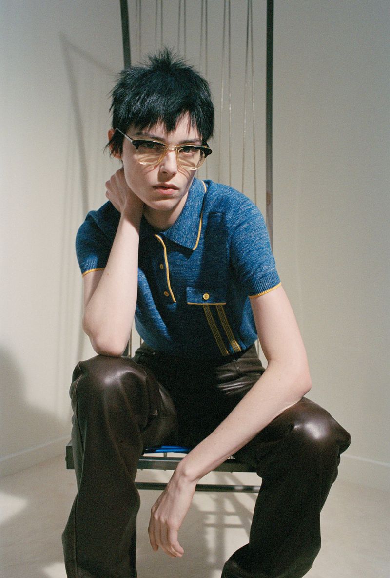 A model with short dark hair is seated in a photo studio, wearing sunglasses, a blue polo shirt, and brown leather pants.