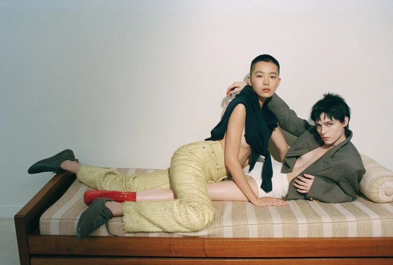 Two fashion models on a striped daybed, one in yellow pants and the other in a brown jacket, showcasing contemporary style and posing.