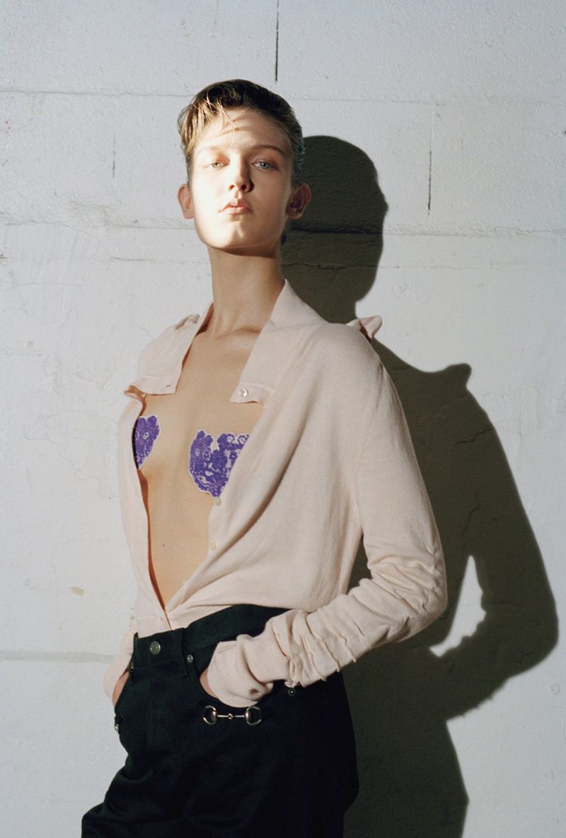 Model with short hair poses in front of a white brick wall, wearing an unbuttoned pale pink shirt, black pants, and purple lace pasties.