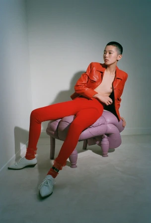 Model with a shaved head wearing a red leather jacket and red pants sitting on a purple tufted stool against a plain grey wall