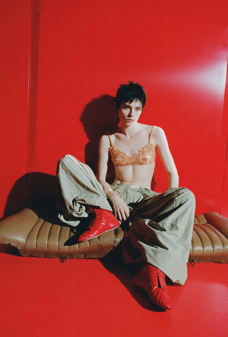 A stylish adult woman with short dark hair poses on a leather couch in red heeled boots against a red backdrop in a fashion shot.