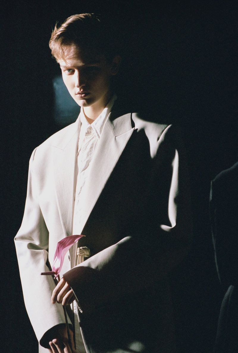 Portrait of a young adult in a light-colored suit and white button-down shirt, standing in a dark environment with shadows and spotlighting.