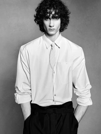 Black and white portrait of a male fashion model with curly hair posing in a button-down shirt and black slacks, against a neutral backdrop.