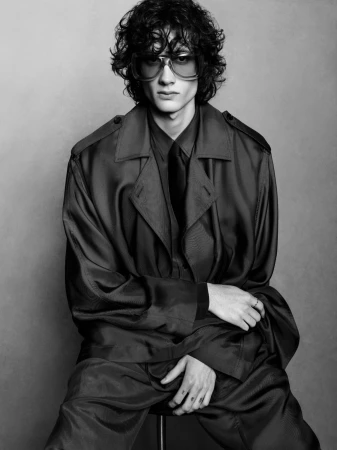 A young man with curly hair wearing oversized sunglasses, a trench coat, and a tie, posed against a plain grey studio backdrop.