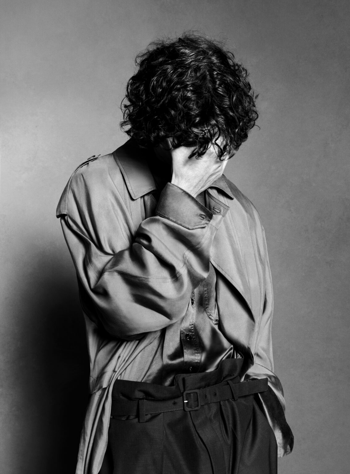 Monochrome portrait of a person with dark curly hair wearing a shiny long-sleeved shirt and dark pants, covering face with hand.