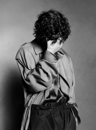 Monochrome portrait of a person with dark curly hair wearing a shiny long-sleeved shirt and dark pants, covering face with hand.