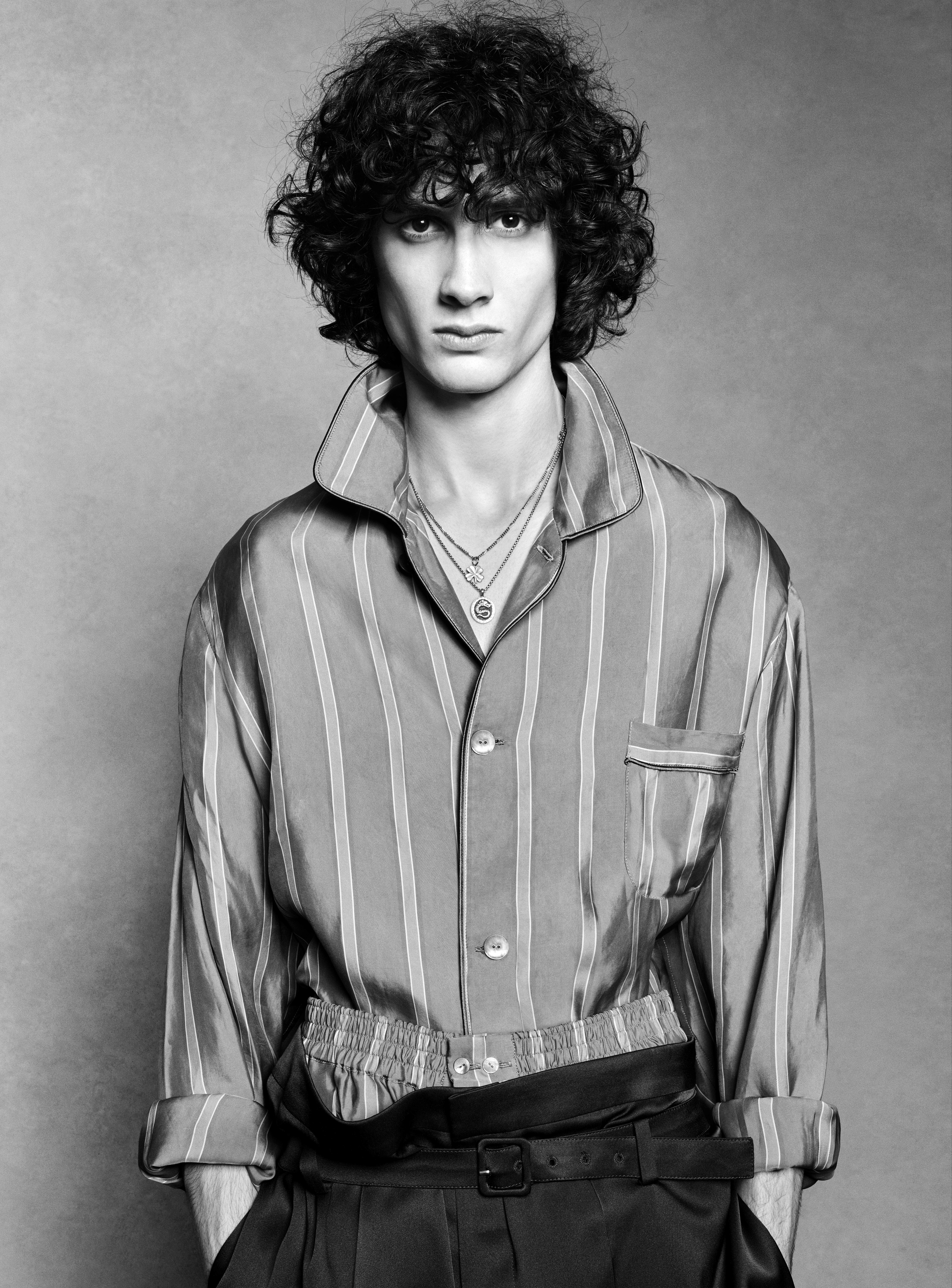 Black and white portrait of a young adult man with curly hair wearing a striped pajama-style shirt with layers of necklaces inside a studio.