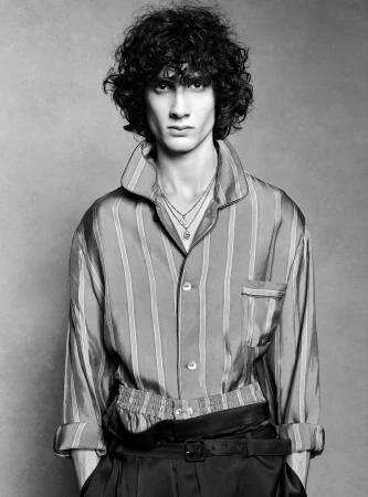 Black and white portrait of a young adult man with curly hair wearing a striped pajama-style shirt with layers of necklaces inside a studio.