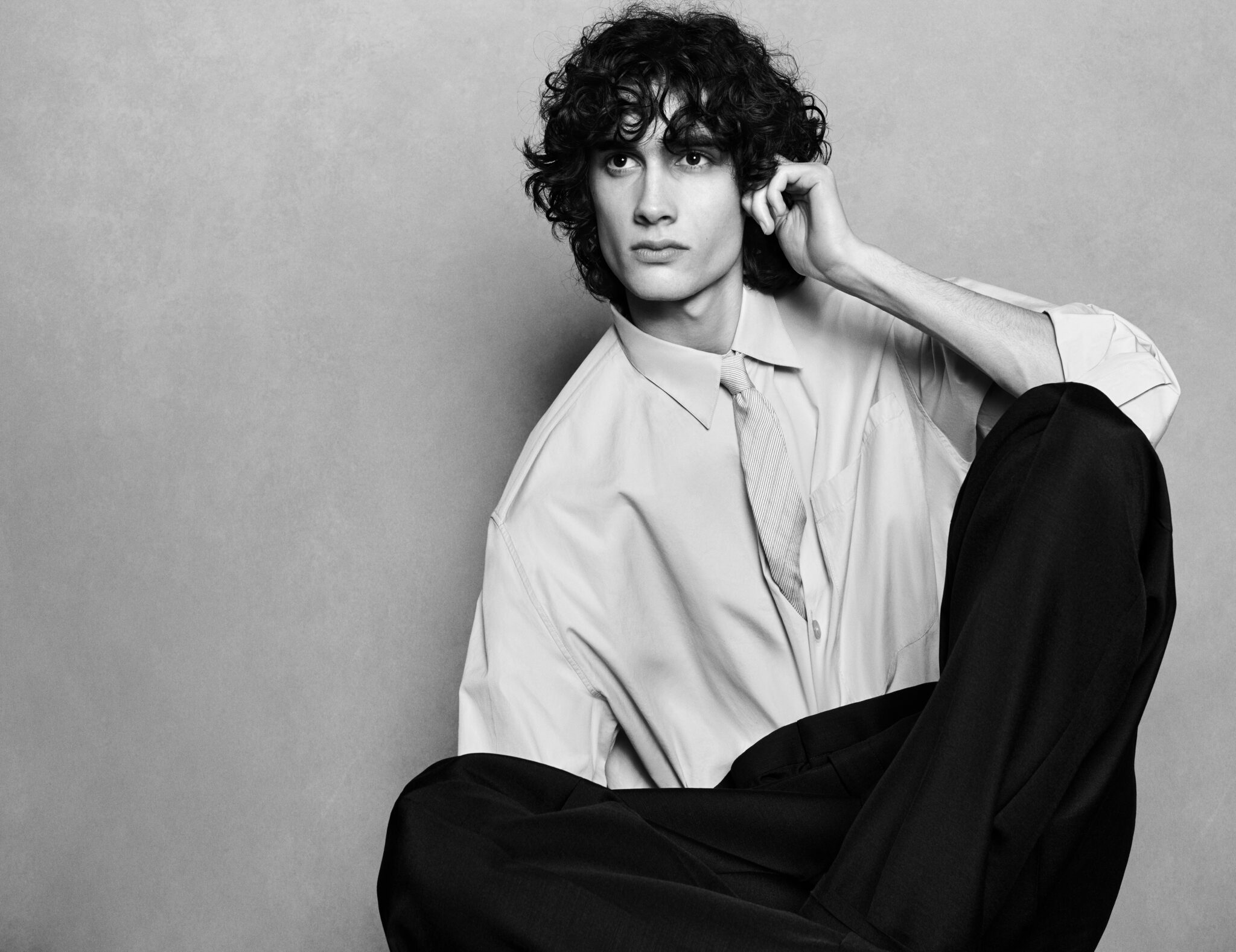Young male model with dark curly hair wearing a light shirt, tie, and dark trousers against a plain grey studio backdrop.