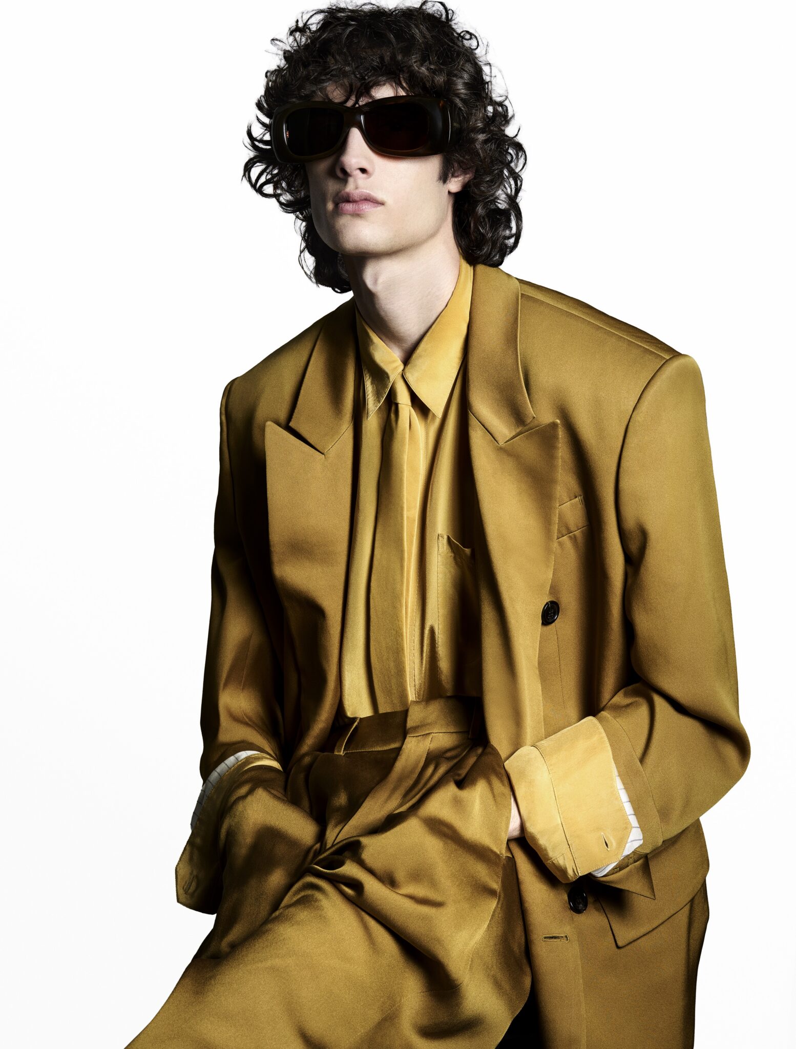 Fashionable man with curly hair, wearing a gold colored suit and sunglasses, poses in a studio with a plain white background.