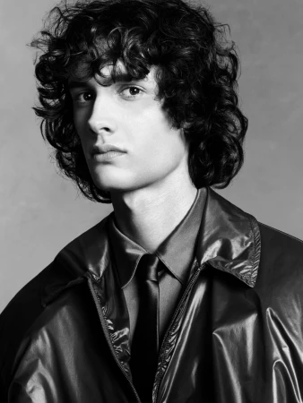 Portrait of young man with dark curly hair wearing a dark collar shirt, tie, and shiny jacket against grey backdrop.