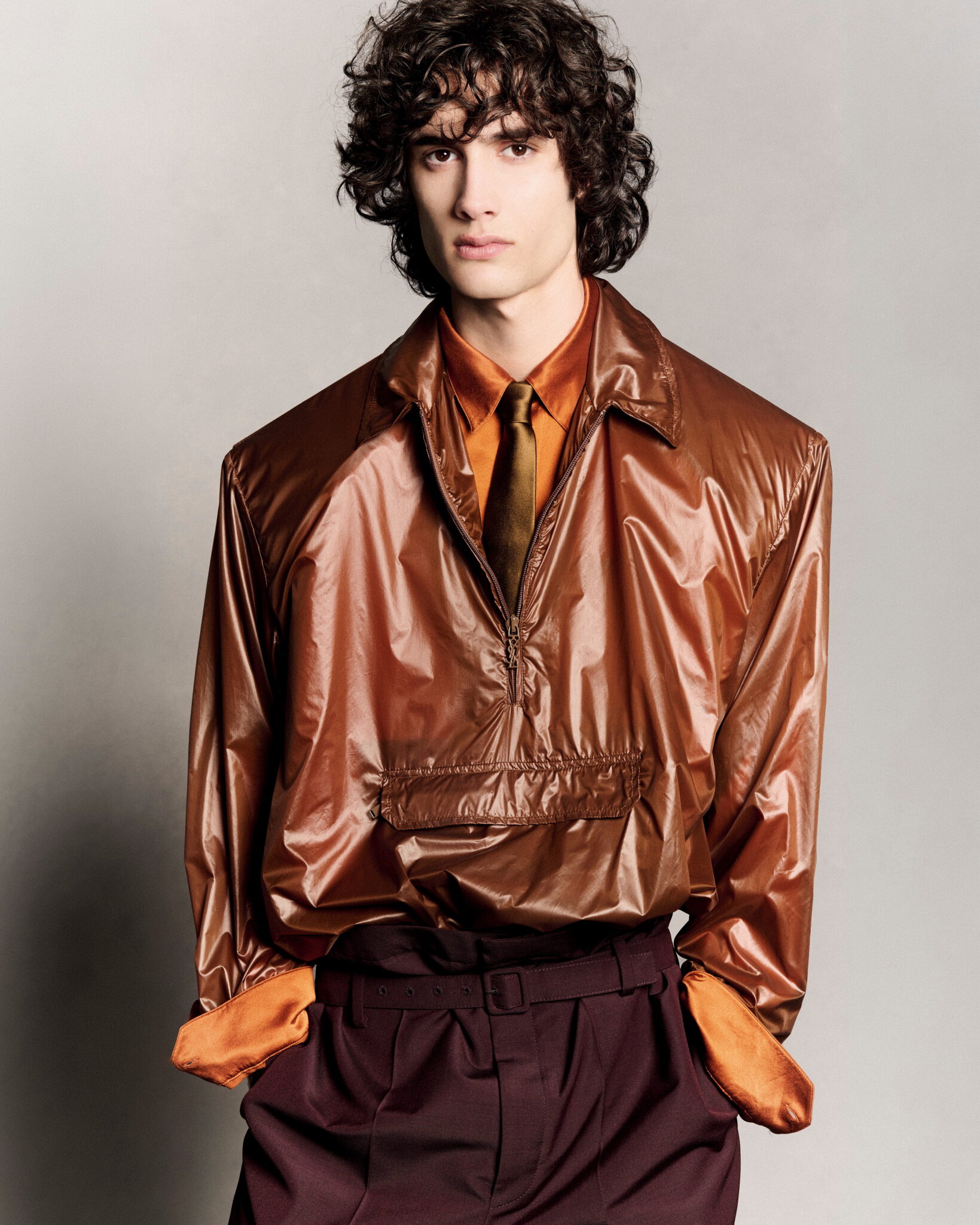 Close-up studio portrait of a young adult male model with curly dark hair, wearing a shiny brown jacket, orange shirt, and maroon pants.