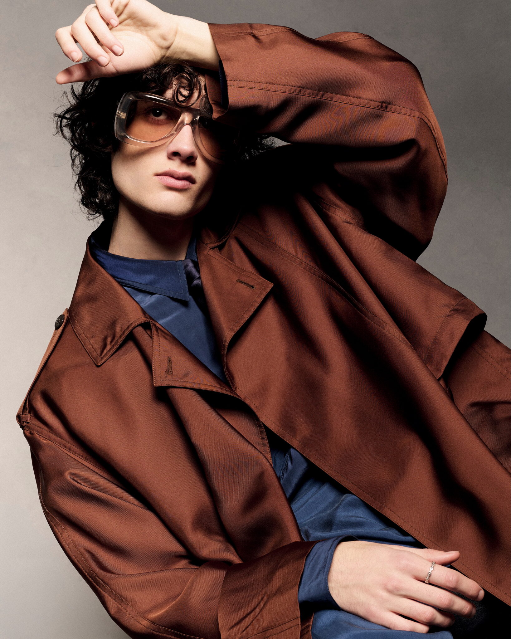 A person with wavy dark hair wearing a glossy brown coat and dark tinted sunglasses, with one arm raised to their forehead.