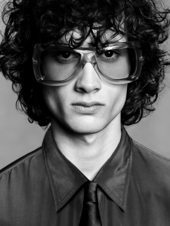 Close-up portrait of a young man with curly dark hair wearing clear framed glasses and a dark shirt with a tie