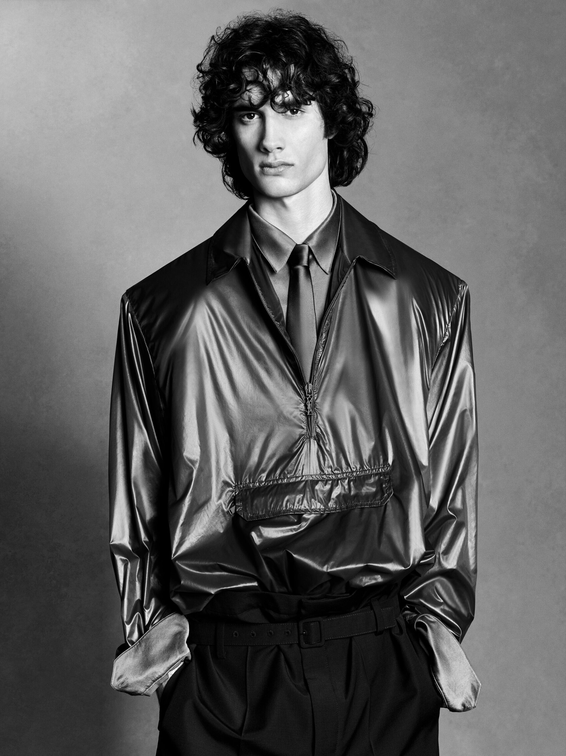 Monochrome portrait of a curly-haired male model in a shiny jacket and dark trousers against a grey backdrop in a studio setting.