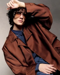 Model in oversized brown trench coat wearing tinted sunglasses and a blue shirt against a neutral background