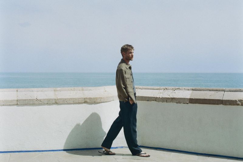 A man in a jacket and pants walking along a terrace with a blue ocean backdrop, captured in a medium shot on a sunny day.