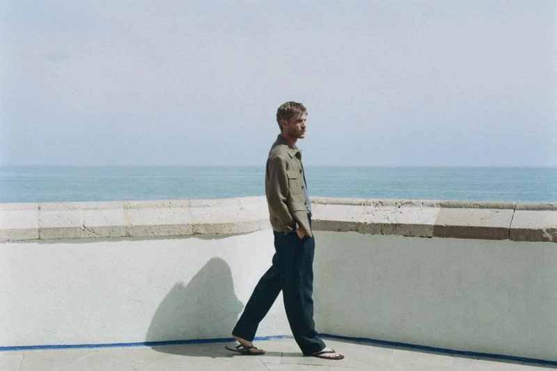 A man in a jacket and pants walking along a terrace with a blue ocean backdrop, captured in a medium shot on a sunny day.